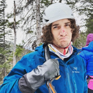 A man looks at the camera with a sad expression. He is wearing ice climbing gear, holding an ice tool over his shoulder, and is bleeding from a cut on the nose. The black Showa Temres gloves he is wearing extend to mid-forearm.
