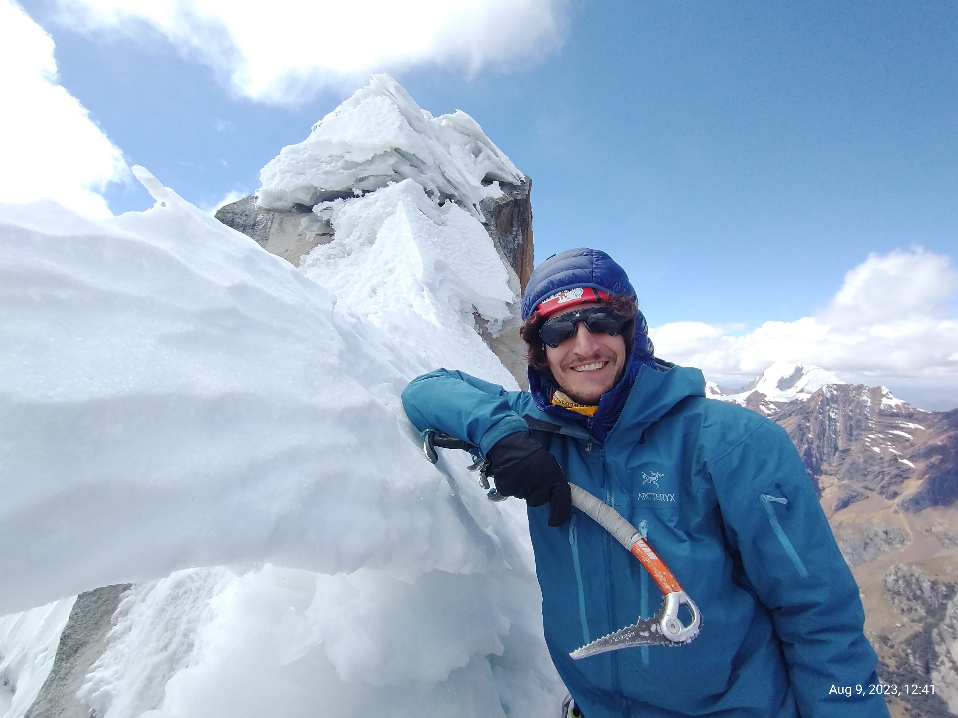 Dan Kay, a male climber, atop a mountain peak in Peru. He rests his right arm on a rib of snow, and holds an aggressive ice tool in his hand. He wears a hardshell jacket, glacier glasses, and a helmet. He is smiling big, on the summit. Blue skies are behind. 