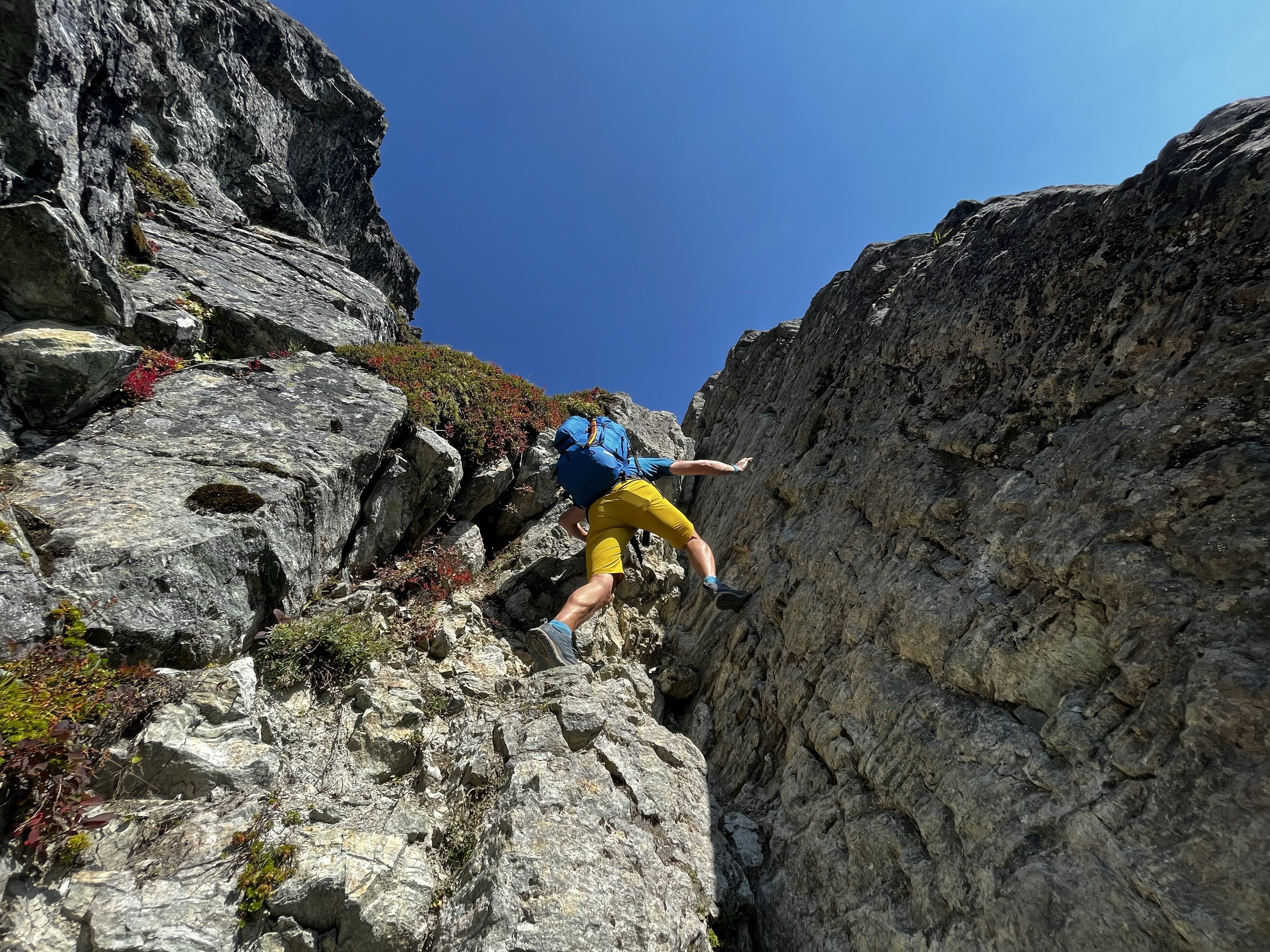 Man climbing rock where two walls meet, in yellow shorts with. abig blue backpack on, no rope. Terrain is not too steep.