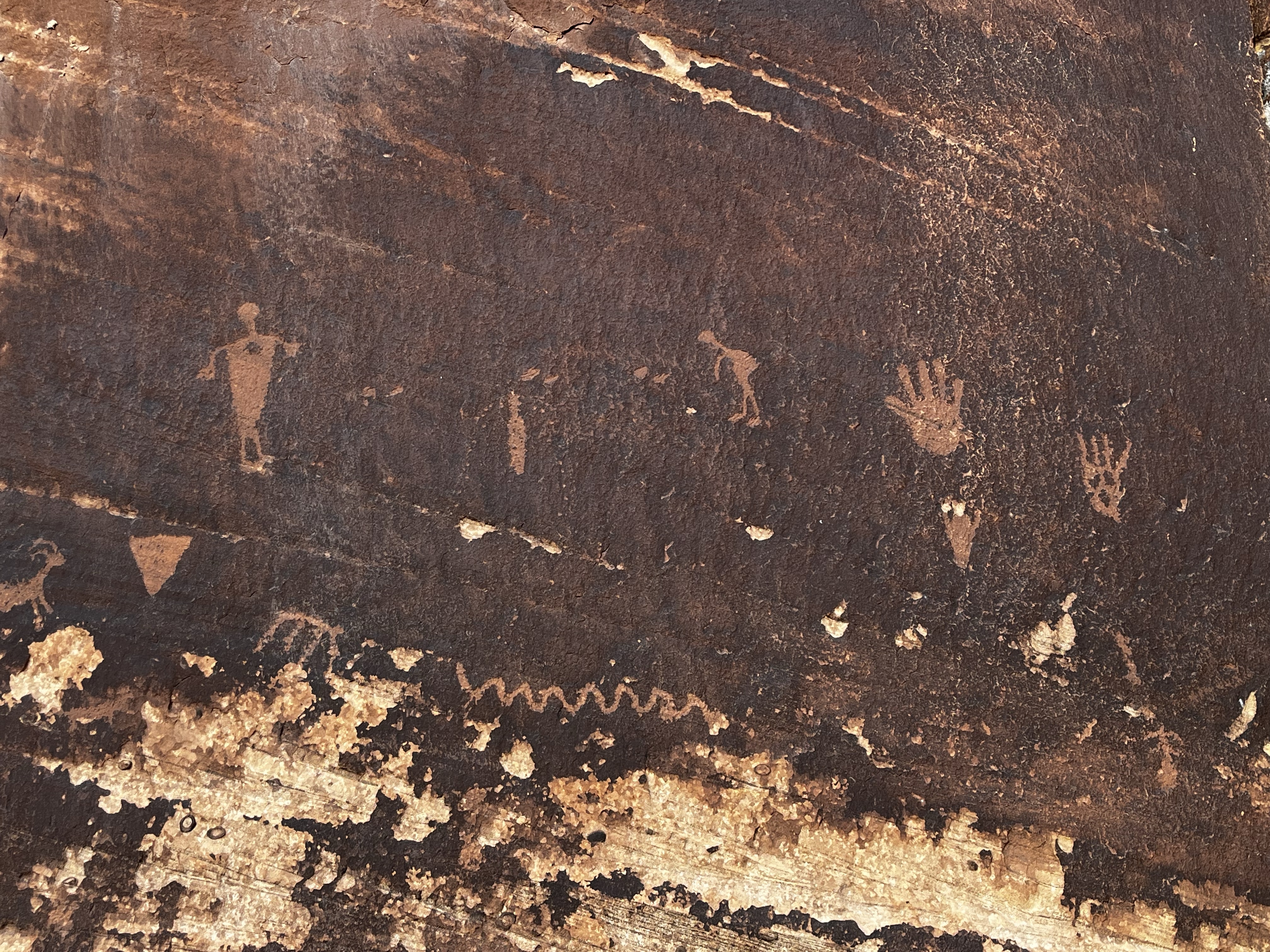 Primitive pictures carved into dark sandstone. Recoghnizeable figures include a person, a Kokopelli, handprints, and depictions of antelope or similar animals. There are also abstract shapes, including a triangle and a snake-like squiggle.