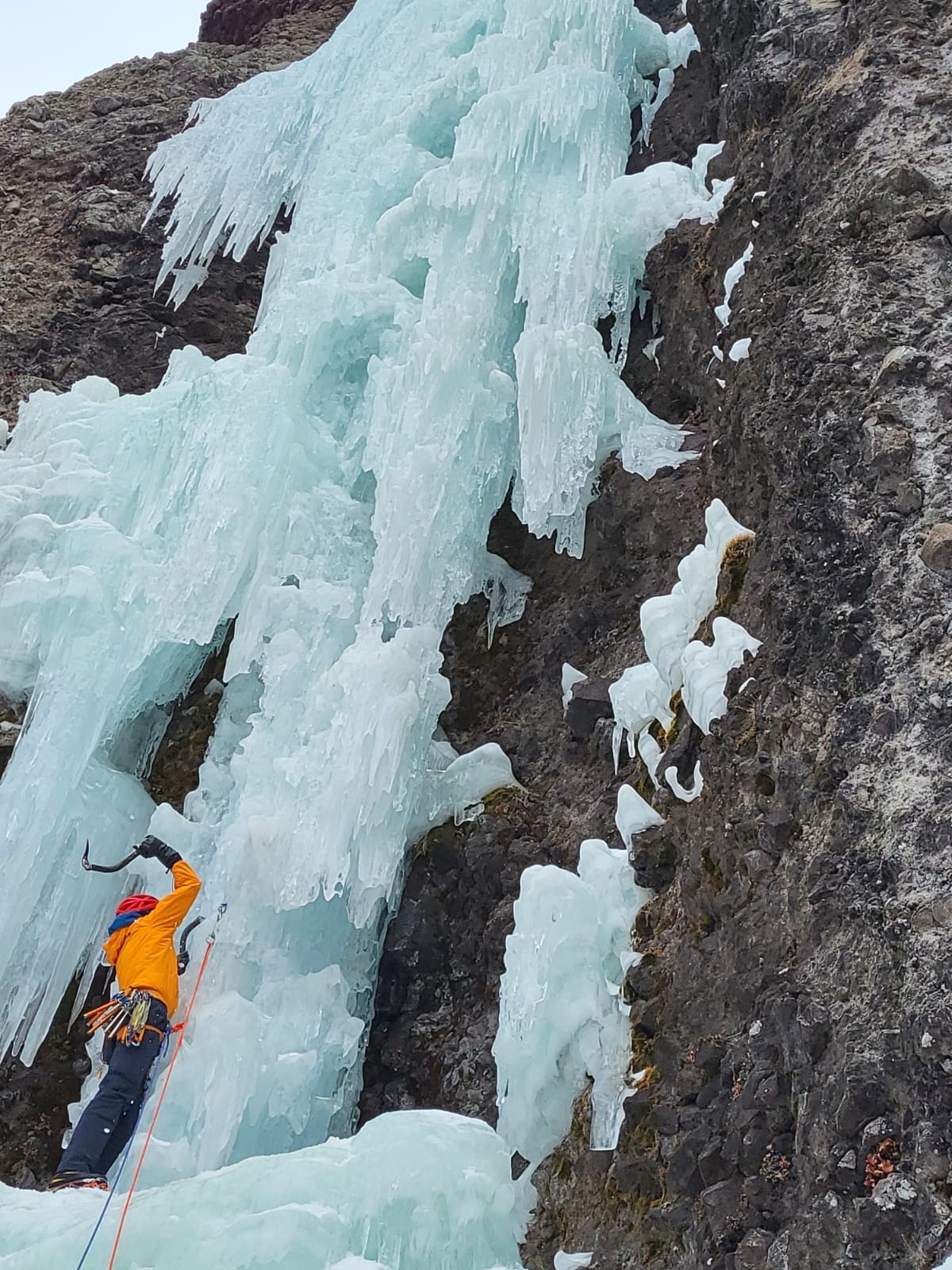 A man stands at the base of a steep pillar of ice, swinging his ice tool to begin the lead. he wears Showa Temres 282-02 Japanese Fishing Gloves, a bright orange jacket, navy blue pants, and a red helmet. An ice screw is placed at his eye level and the rope is attached to provide fall protection as he begins the climb.